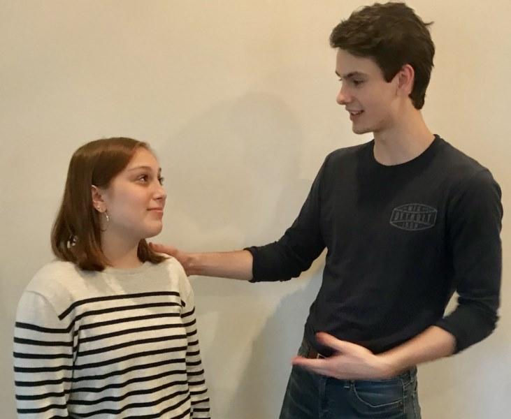 Two students acting out a role play. A tall student has his hand on the shoulder of a girl. He is reassuring her, but she looks skeptical.