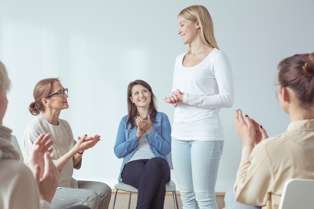 Four women sitting in a circle and clapping as a fifth woman stands and shares some achievement